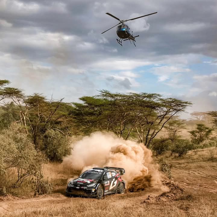 Adrenaline-Fueled Kenyan Safari Rally: A Spectacle of Speed and Dust A rally car kicks up a cloud of dust as it speeds through the Kenyan savannah, with a helicopter capturing the thrilling action above.