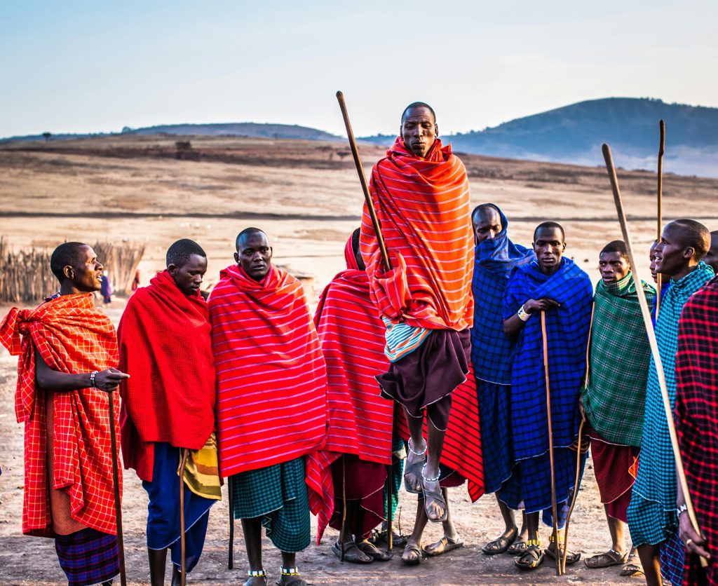 A group of Maasai men in vibrant traditional attire performing a cultural jumping dance outdoors.
